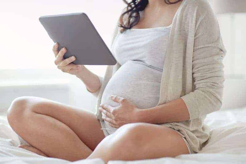 Pregnant woman sitting on a bed and looking at a tablet while resting a hand on her stomach.