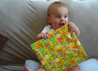 Excited baby holding a yellow birthday gift while sitting on a couch.