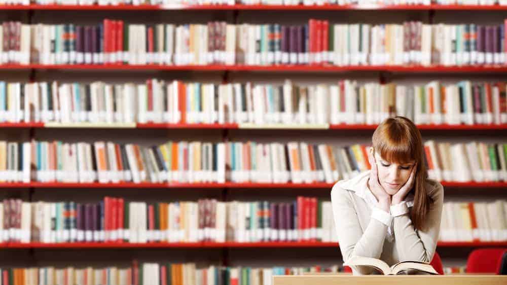 A woman with auburn hair reads a book in front of multiple rows of bookshelves in a library.