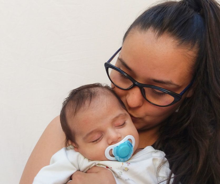 Close-up of a woman in glasses holding and kissing a sleeping infant with a blue pacifier.