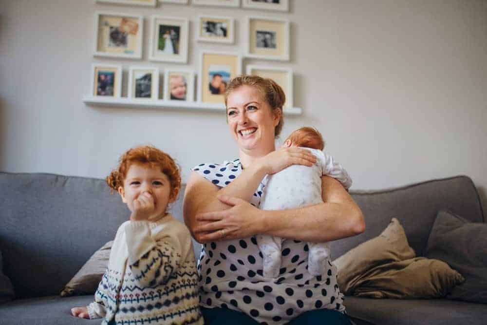 A smiling woman sitting on a sofa holds a baby while a young child with red hair sits beside her.