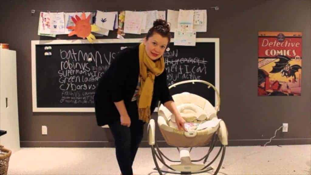 A woman in a yellow scarf demonstrates a Graco Glider LX baby swing in a room with a chalkboard and Batman poster.