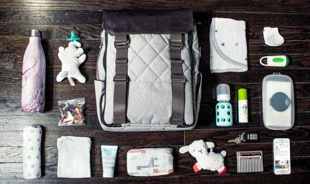 A grey diaper bag backpack surrounded by baby essentials like bottles, diapers, wipes, toys, and clothes on a wood floor.