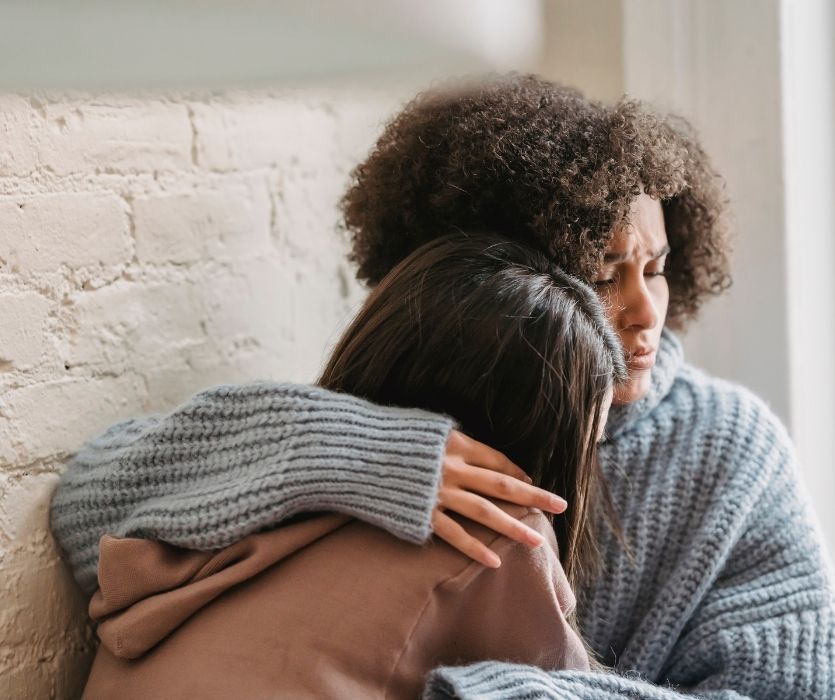 A woman with a sad expression embraces another person in a supportive hug against a white brick wall.