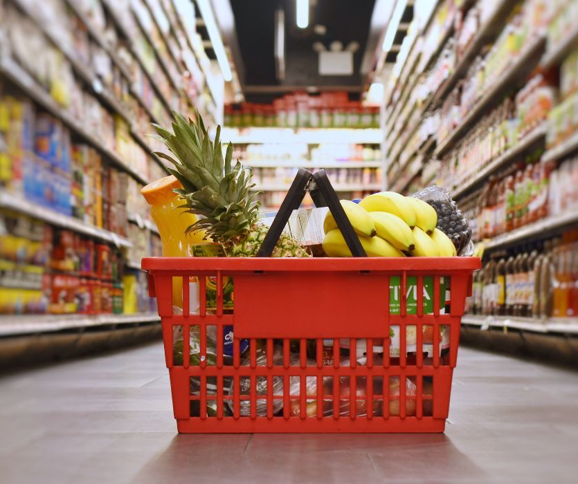 Red grocery basket filled with a pineapple, bananas, and orange juice sitting in a grocery store aisle.