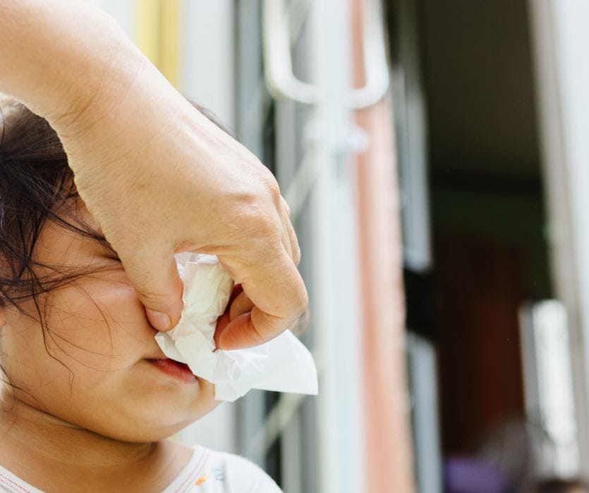 An adult hand holds a white tissue to a young child's nose to help them blow it.