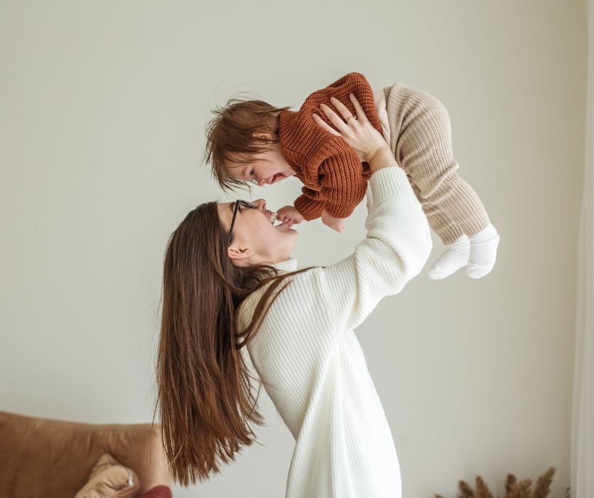 A woman in a white sweater smiling and lifting a laughing baby in a brown knit sweater into the air.