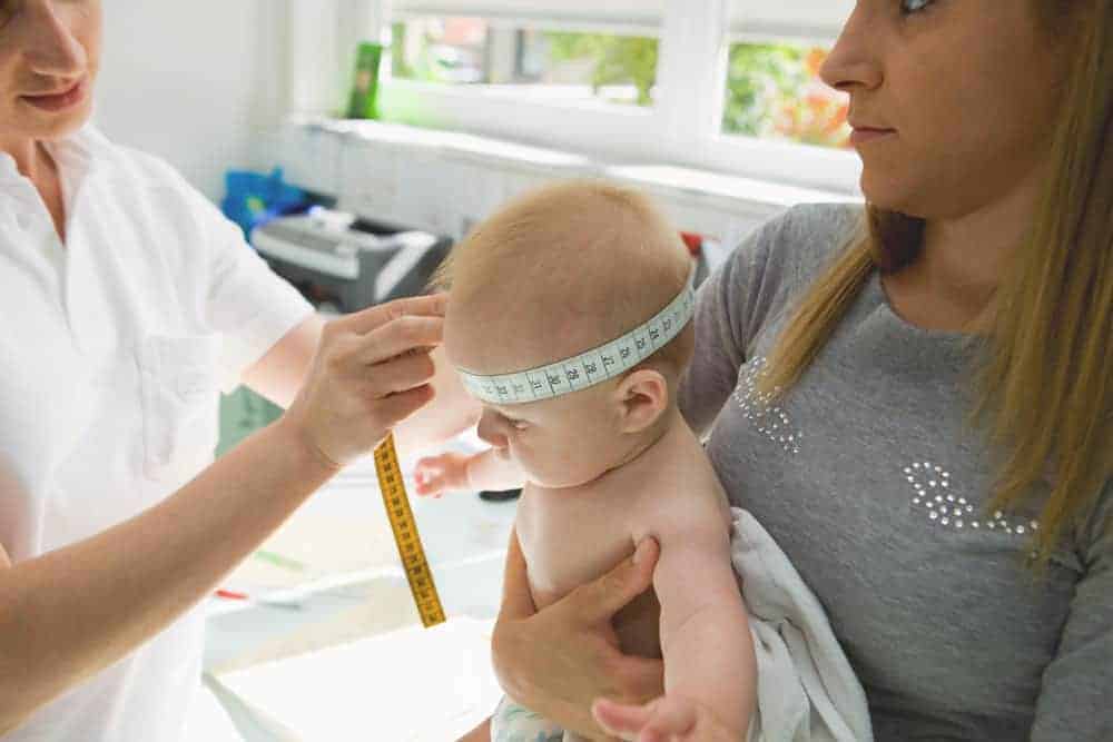 A healthcare professional uses a measuring tape to check a baby's head circumference while a woman holds the child.