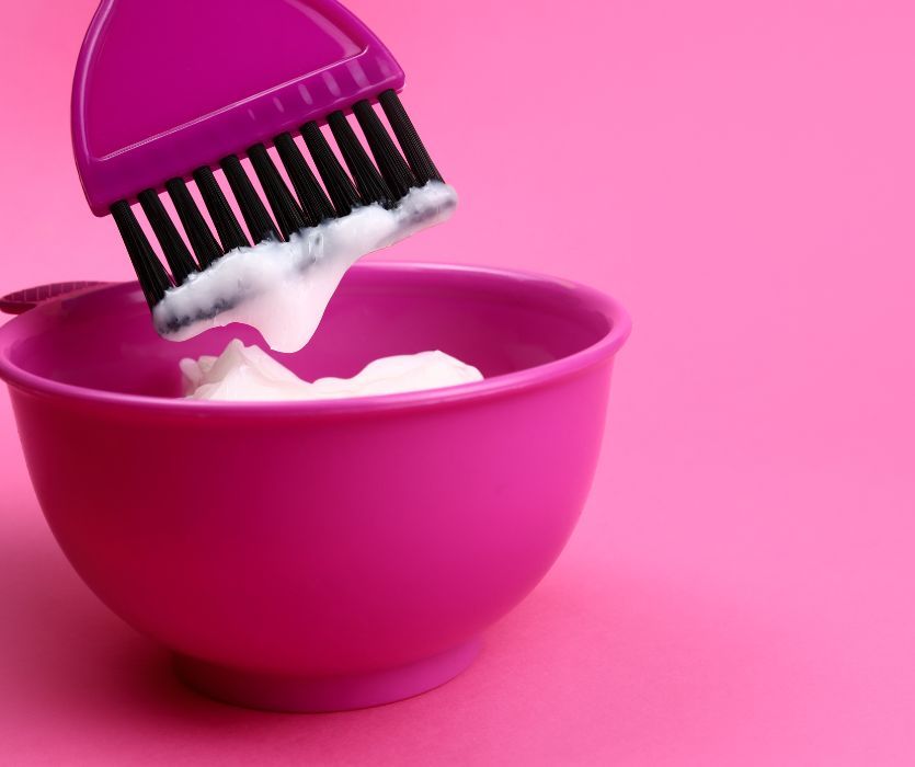 A purple hair dye brush with white cream held over a matching purple mixing bowl against a pink background.