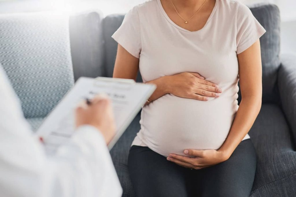 Pregnant person holding their belly while a healthcare provider in a white coat takes notes on a clipboard.