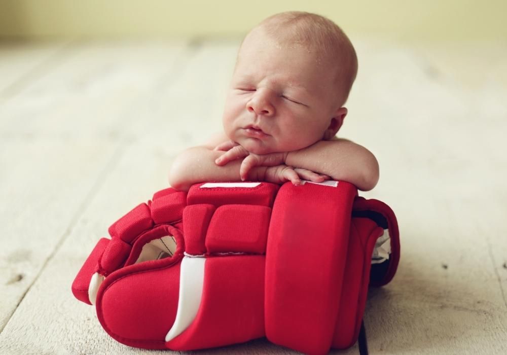 A sleeping newborn baby rests their head on their folded hands atop a red hockey glove.