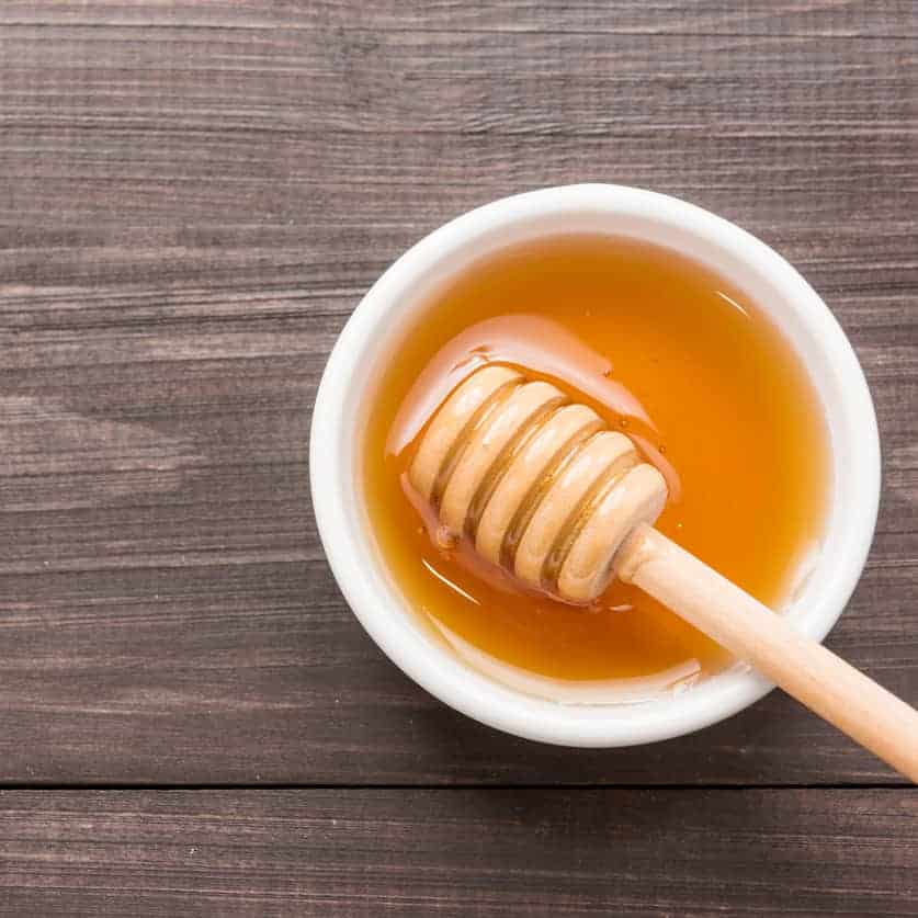 Top-down view of a wooden honey dipper resting in a small white bowl of golden honey on a dark wooden table.