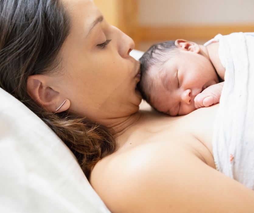 A mother lies down holding her newborn baby skin-to-skin and kisses the infant's forehead.