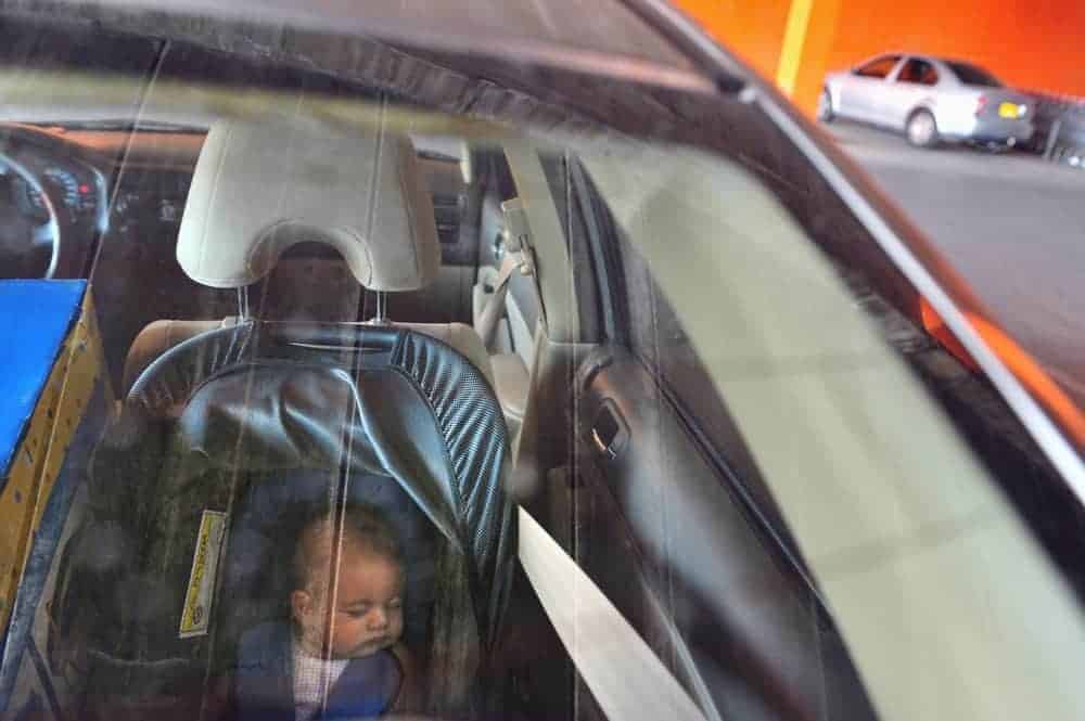 An infant sleeping in a car seat inside a parked vehicle, seen through a window.