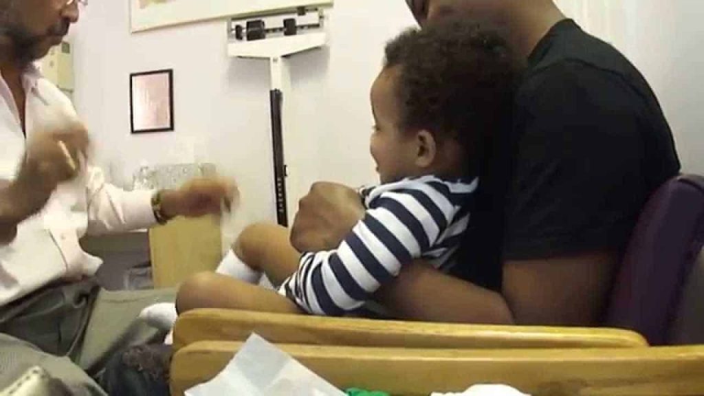 A doctor uses sleight of hand to distract a toddler sitting in a parent's lap during a checkup.