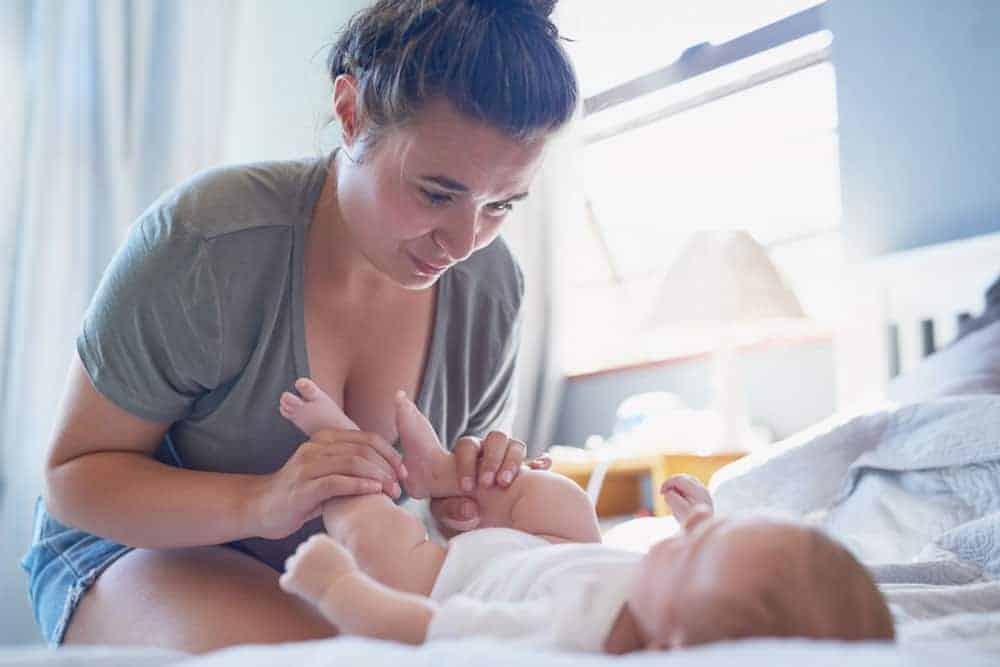 Tired woman looking down while changing a baby's diaper on a bed.