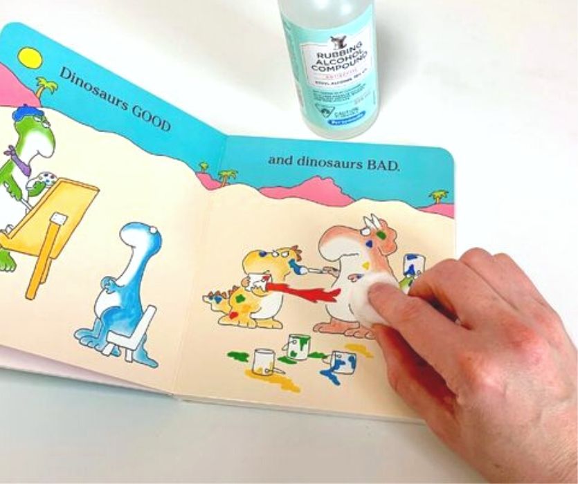 A hand wipes a dinosaur board book with a cotton ball near a bottle of rubbing alcohol.
