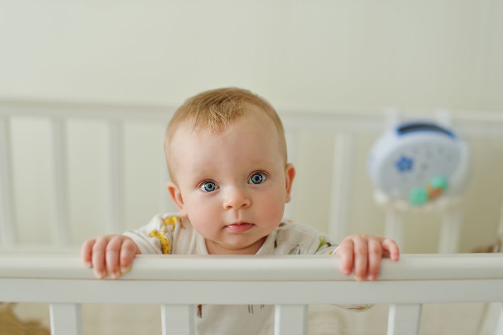 Blue-eyed baby standing in a white crib, holding onto the railing and looking forward.