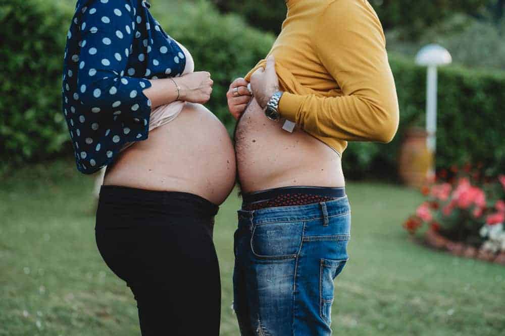 A pregnant woman and a man standing face-to-face, exposing and comparing their bellies in a garden.