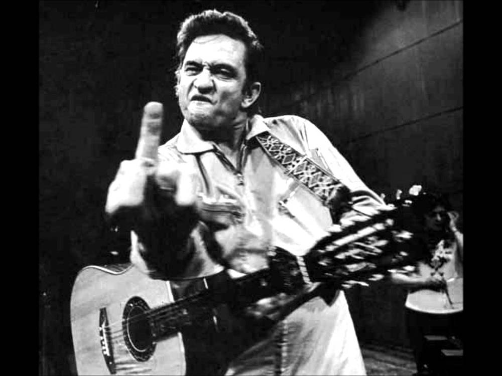 Black and white photo of Johnny Cash scowling and gesturing with his middle finger while holding an acoustic guitar.