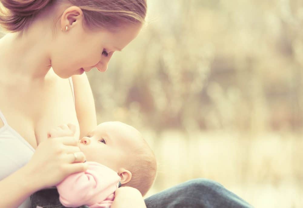 A woman looking down at a baby as she breastfeeds outdoors against a blurred background.