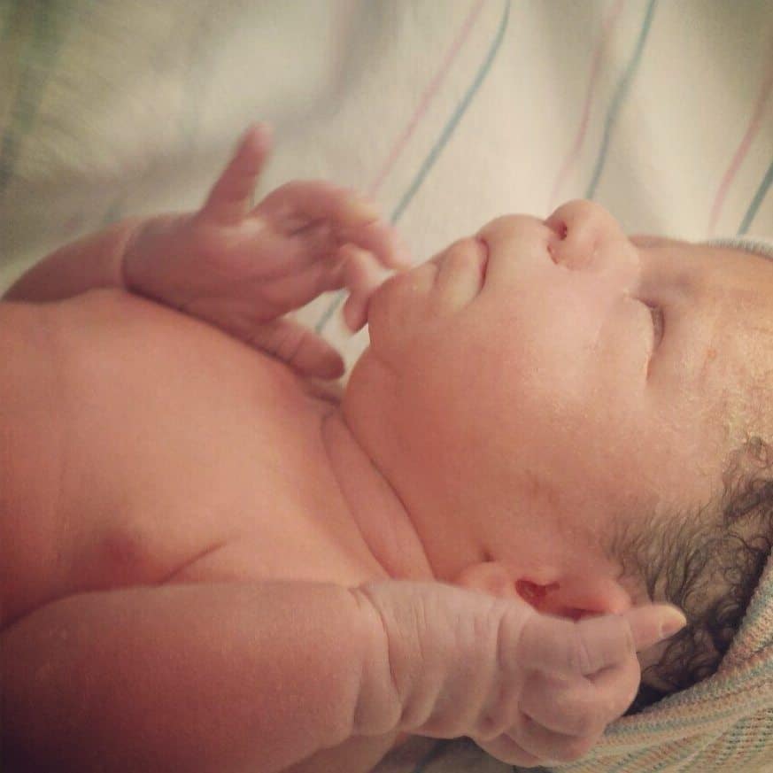 Close-up of a newborn baby lying down with eyes closed, hands near their face.