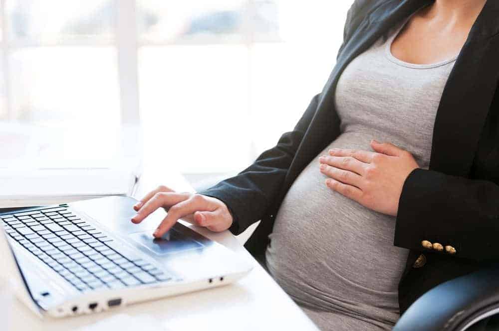 Close-up of a pregnant person in a blazer working on a laptop at a white desk.