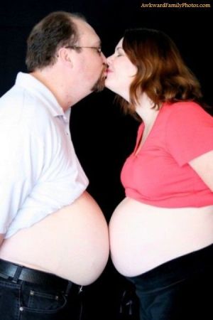 A man and a pregnant woman kissing while their bare stomachs touch against a black background.