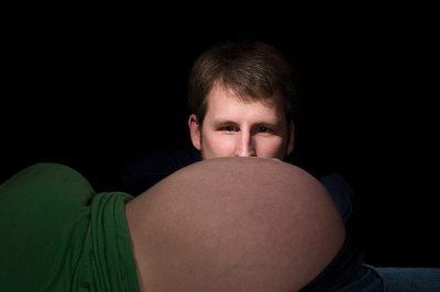 A man peeking over a pregnant woman's bare belly against a black background.