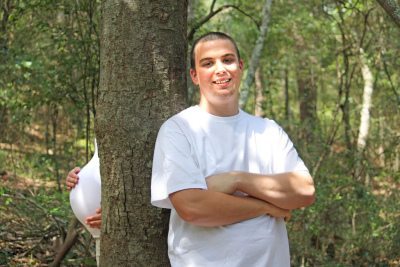 Smiling man with arms crossed leaning against a tree in a forest next to a pregnant belly peeking from behind the trunk.
