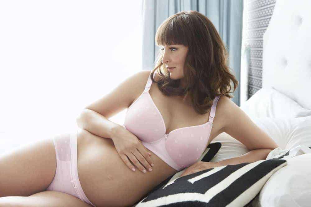 Pregnant woman with brown hair and bangs wearing a light pink polka dot bra and panty set while lounging on a bed.