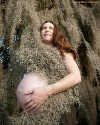 A pregnant woman with long red hair posing outdoors while draped in thick Spanish moss.