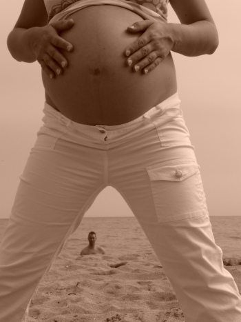 Sepia photo of a pregnant person holding her belly on a beach, with a small person appearing between her legs.