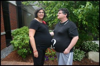 A pregnant woman and a man in a shirt with a question mark on his stomach standing together outdoors.