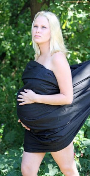 A pregnant blonde woman stands in a garden, her belly wrapped in a long black fabric.