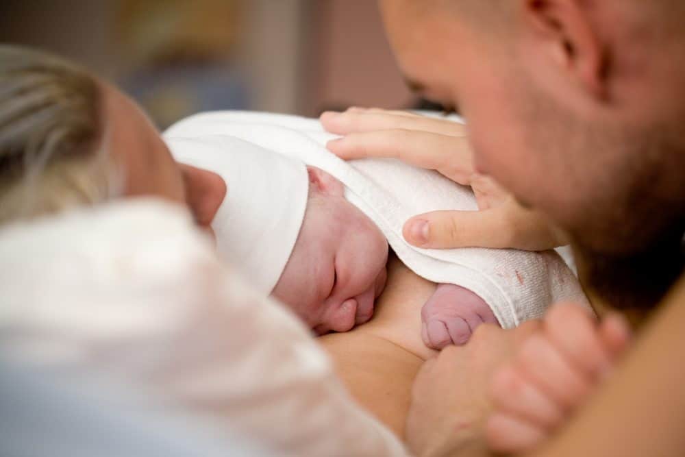 A newborn baby in a white hat rests on a mother's chest while a father looks on and touches the baby's shoulder.