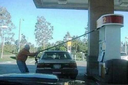 A man pulls a stretched gas pump hose across the back of a sedan at a gas station.