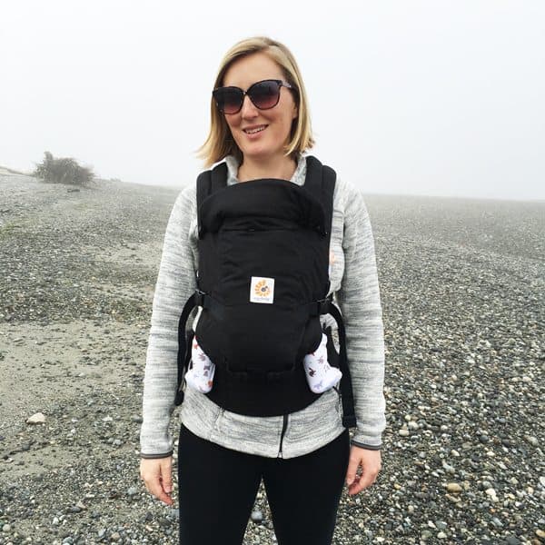 Woman wearing sunglasses and a black Ergobaby carrier with a baby inside on a pebble beach.