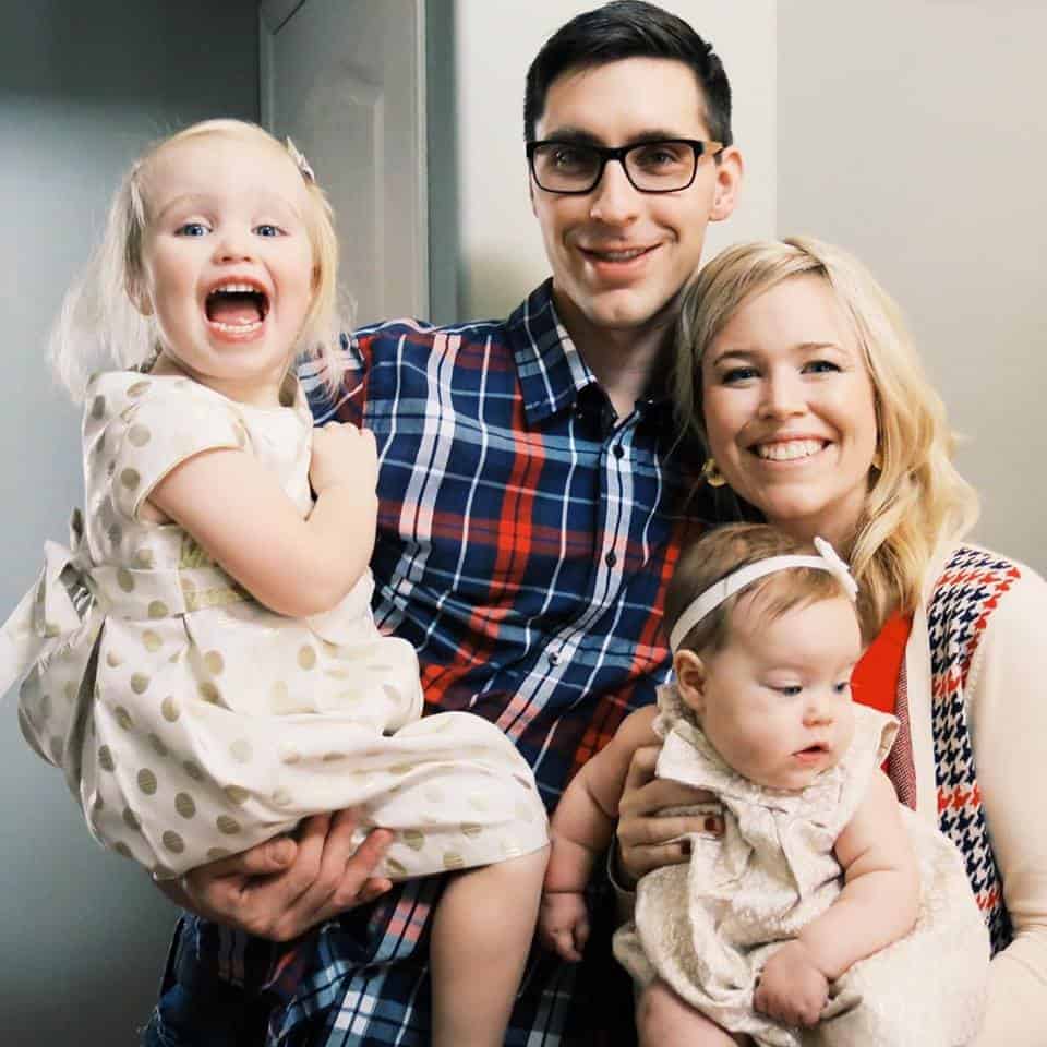 A smiling family of four, including a man in a plaid shirt and a woman holding two young daughters.