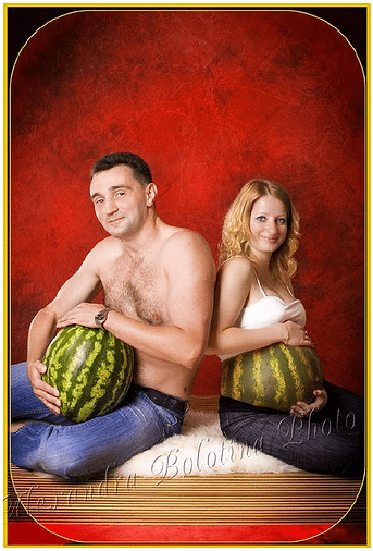 Shirtless man holding a watermelon sits back-to-back with a pregnant woman holding a watermelon over her belly.