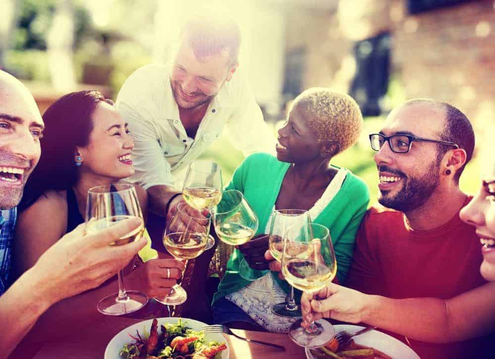 Diverse group of friends smiling and clinking wine glasses over a meal outdoors.