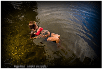 A pregnant woman with a tattooed belly and red top floats in dark, rippling water.