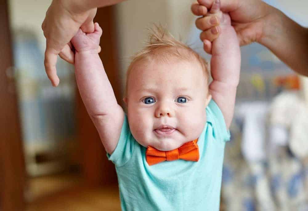 Baby wearing a light blue shirt and orange bow tie held by the hands while standing.