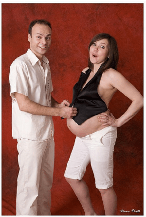 Man in white shirt buttoning a black vest over a pregnant woman's belly against a red backdrop.