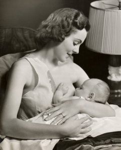 A vintage black and white photograph of a woman breastfeeding an infant while sitting next to a lamp.