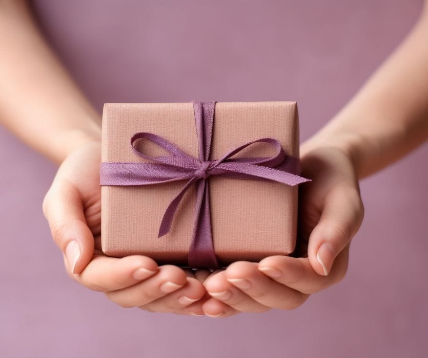 Hands holding a small pink gift box tied with a purple ribbon.