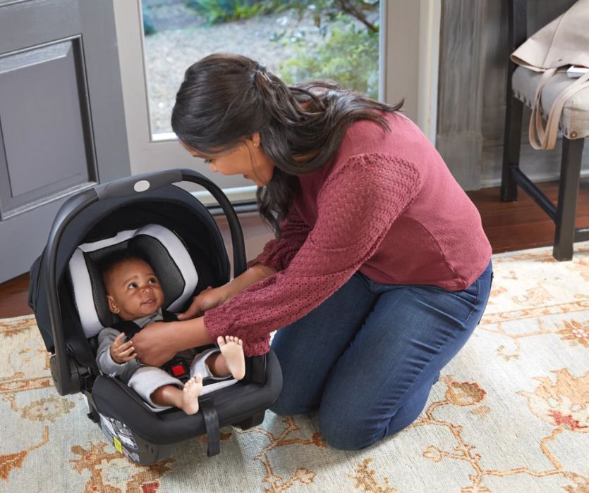 A woman buckles a smiling infant into a black and white rear-facing infant car seat on a rug indoors.