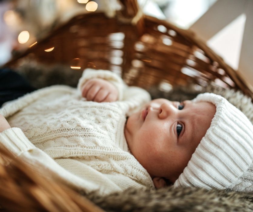 An infant wearing a white cable knit sweater and beanie lying in a wicker basket with soft Christmas lights.