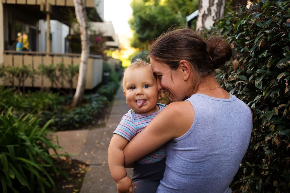 A woman holds a smiling toddler sticking his tongue out while standing on a path outside.