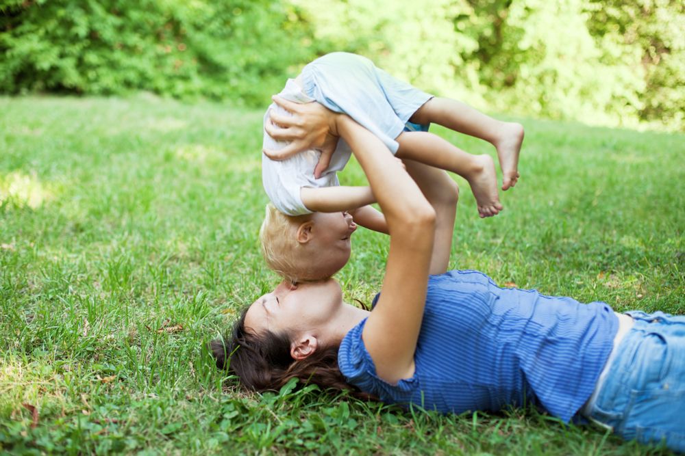 A woman lying on the grass holds a toddler upside down as the child playfully puts their mouth over hers.